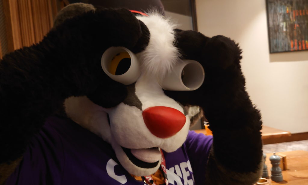 A photo of Brok the Badger (Confuzzled's mascot) fursuit, holding two glasses to their eyes
