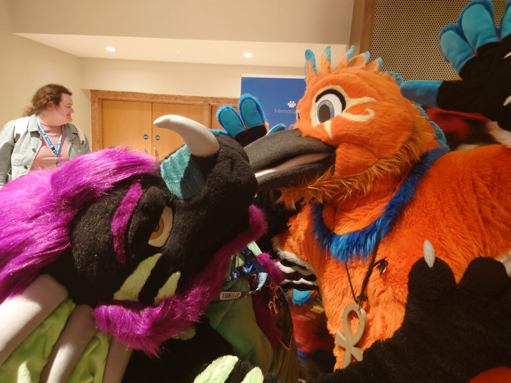 A photo of Crome, a green and black dragon with purple hair, and Regdeh, a orange hoopoe. Regdeh's beak is in Cromes maw for, reasons?
