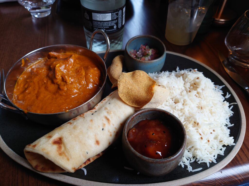 A photo of a Chicken Masala curry in a metal bowl, on a plate with rice, a small bowl of mango chutney, a small bowl of onions, a naan that has been rolled up to save space and some small popadom crisps