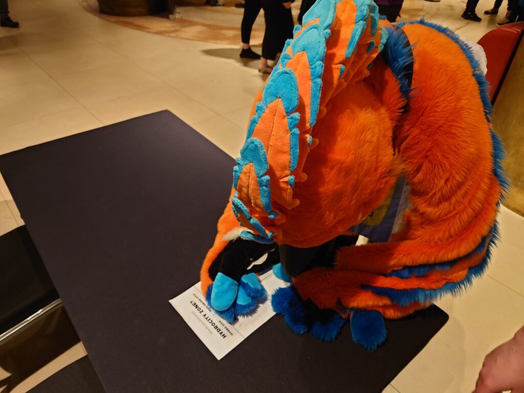 A photo of Regdeh, an orange and blue hoopoe fursuiter, casting their vote in the Hydrocity pronunciation debate.